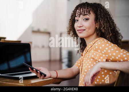 Jeune femme indépendante tenant un smartphone assis près d'un ordinateur portable dans un café Banque D'Images