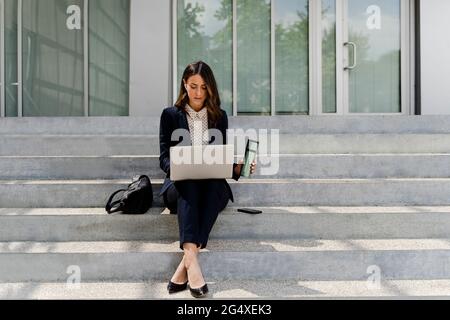 Femme d'affaires tenant une tasse de voyage à l'aide d'un ordinateur portable tout en étant assise dans un escalier Banque D'Images