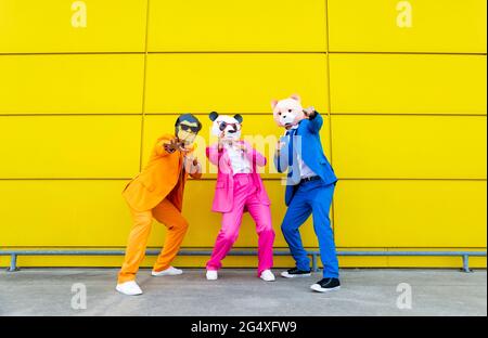 Trois adultes portant des costumes vibrants et des masques pour animaux se posant ensemble devant le mur jaune Banque D'Images