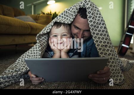 Un père et un fils souriants avec une tablette numérique couchée sous une couverture dans le salon Banque D'Images