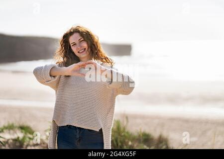 Jeune femme souriante montrant la forme du cœur à la plage Banque D'Images