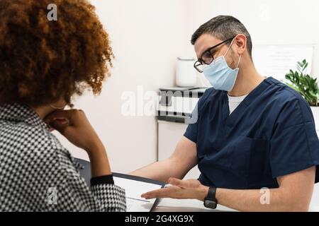 Dentiste de sexe masculin portant un masque facial de protection discutant de la carte dentaire avec une patiente à la clinique Banque D'Images