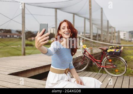 Femme prenant le selfie à travers le smartphone tout en étant assise sur la jetée Banque D'Images