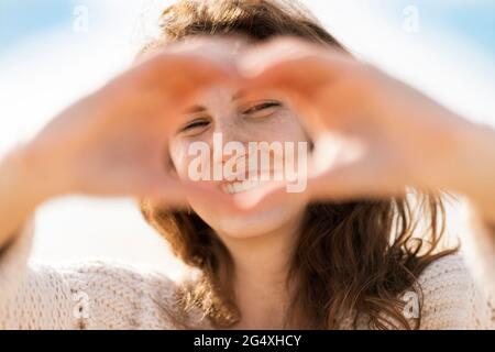 Bonne jeune femme regardant à travers la forme de coeur faite à la main sur la plage pendant la journée ensoleillée Banque D'Images