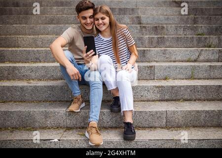 Couple souriant prenant le selfie par téléphone portable tout en étant assis sur des marches Banque D'Images