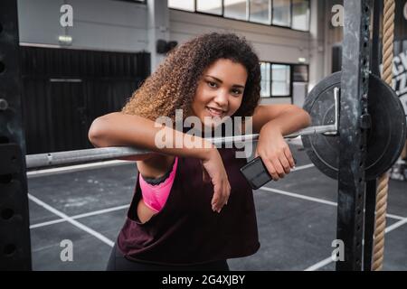 Athlète féminine souriante avec téléphone mobile appuyé sur la barbell à la salle de gym Banque D'Images