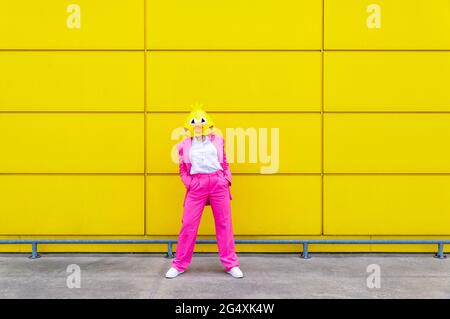 Femme portant un costume rose vif et un masque d'oiseau debout avec les mains dans les poches devant le mur jaune Banque D'Images