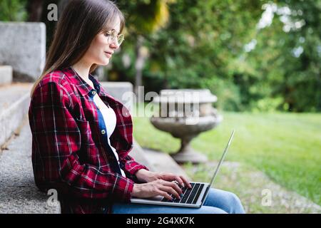 Jeune femme portant des lunettes à l'aide d'un ordinateur portable dans un parc public Banque D'Images