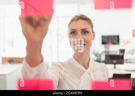 Belle femme entrepreneur souriante écrivant sur des notes adhésives vues à travers le verre dans le bureau Banque D'Images