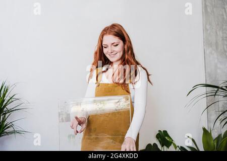 Jeune femme souriante touchant un appareil en verre devant le mur dans la salle de séjour Banque D'Images