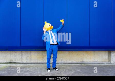 Homme portant un costume bleu vif et un masque d'oiseau prenant le selfie de smartphone devant le mur bleu Banque D'Images