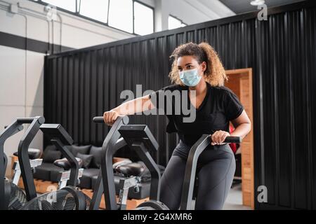 Femme active portant un masque de protection pendant le cyclisme en salle de gym pendant le COVID-19 Banque D'Images