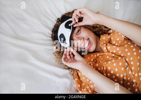 Jeune femme souriante portant un masque oculaire en étant couché sur le lit à la maison Banque D'Images