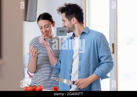 Petit ami souriant nourrissant de la tomate à la petite amie dans la cuisine à la maison Banque D'Images