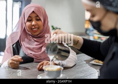 Femme souriante attendant pendant que la serveuse verse du lait dans une tasse au café-restaurant Banque D'Images