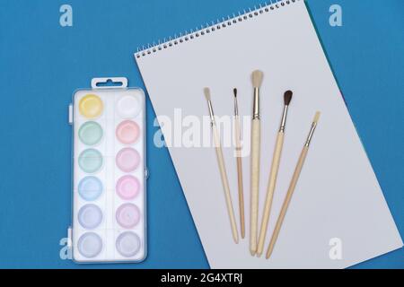 un ensemble de pinceaux, un carnet de croquis et des aquarelles lumineuses sur fond bleu. le concept de la créativité Banque D'Images