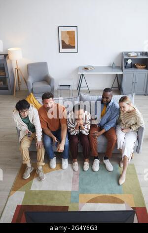 Vue verticale en grand angle de divers groupes d'amis regardant la télévision à la maison tout en étant assis sur le canapé, dans l'espace de copie Banque D'Images