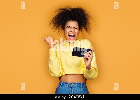 Une fille heureuse a serré le poing dans le geste de victoire, la victoire dans le jeu. Photo d'une fille afro-américaine utilisant un smartphone sur fond jaune Banque D'Images