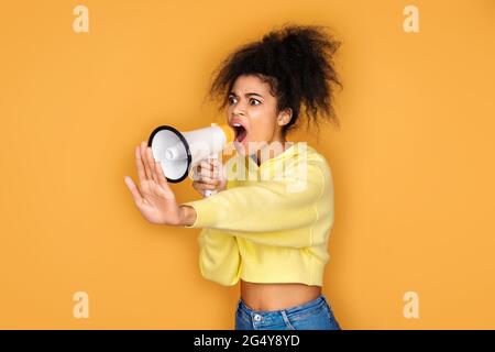 Une fille crie au mégaphone et montre un geste d'arrêt. Photo d'une fille afro-américaine sur fond jaune Banque D'Images