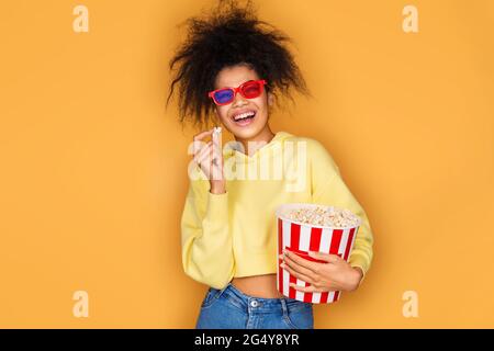 Jeune fille regardant la télévision avec boîte à pop-corn. Photo d'une fille afro-américaine sur fond jaune Banque D'Images