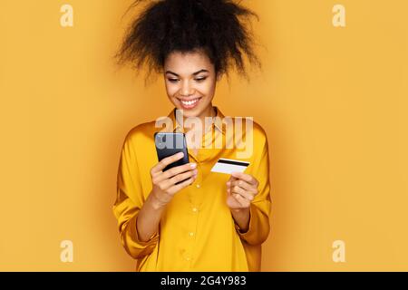 Jeune fille fait un paiement, en utilisant une carte de crédit et un smartphone. Photo d'une fille afro-américaine sur fond jaune Banque D'Images