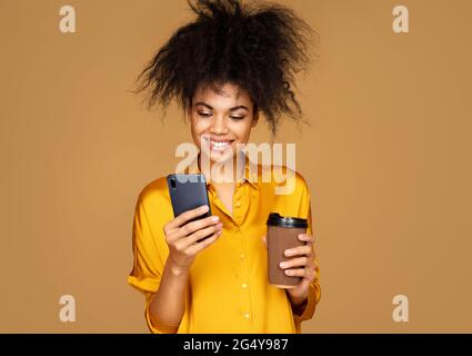 Une fille boit du café et utilise le téléphone, recherche un nouveau message. Photo d'une fille afro-américaine sur fond beige Banque D'Images