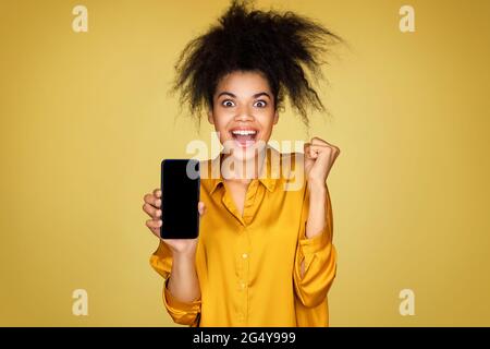 Une fille excitée montre un smartphone et des clenches poing, se sent trop joyeuse après avoir gagné ou de bonnes nouvelles. Photo d'une fille afro-américaine sur fond jaune Banque D'Images