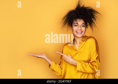 La jeune fille souriante pointe vers l'extérieur avec l'index, s'affiche sur l'espace de copie pour votre texte. Photo d'une fille afro-américaine sur fond jaune Banque D'Images