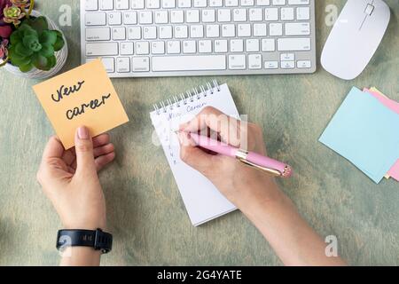 Business and Finance concept. Les mains des femmes tiennent note avec des mots NOUVELLE CARRIÈRE et écriture dans le bloc-notes sur une table en bois. Vue de dessus du bureau avec clavier, cactus et mause. Pose à plat Banque D'Images