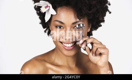 Femme afro-américaine souriante avec une fleur dans les cheveux en utilisant le boucleur de cils isolé sur le gris Banque D'Images