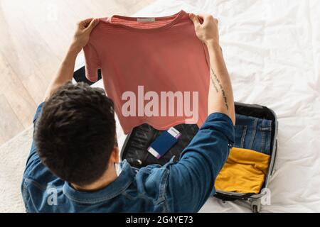 Vue en hauteur de l'homme tenant un t-shirt près d'une valise avec passeport sur le lit Banque D'Images