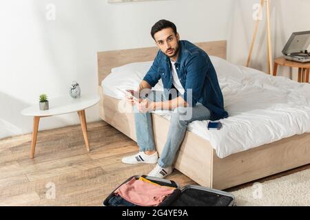 Jeune voyageur avec tablette numérique regardant l'appareil photo près de la valise et passeport dans la chambre Banque D'Images