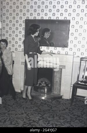 1957, historique, lors d'une fête à l'hôtel Victoria, Leeds, une jeune femme portant une élégante robe debout près de la cheminée donnant sur un miroir. Banque D'Images