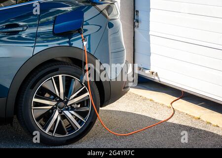 Une voiture électrique hybride garée devant la porte de garage de la maison est branchée avec un câble de charge pour recharger la batterie. Banque D'Images
