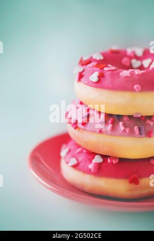 De délicieux beignets roses, mignons et brillants, avec des arrosettes sous forme de cœurs, sont couchés sur une soucoupe rouge. Un cadeau romantique et appétissant pour la Saint-Valentin Banque D'Images