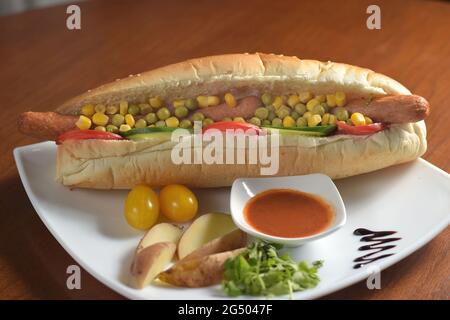 Long hot dog avec deux saucisses, petits pois verts et maïs, avec des tranches de tomates et de concombres, sur une assiette blanche avec sauce, pommes de terre et toma de cerise Banque D'Images