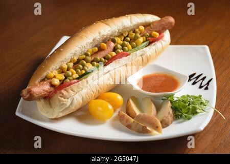 Long hot dog avec deux saucisses, petits pois verts et maïs, avec des tranches de tomates et de concombres, sur une assiette blanche avec sauce, pommes de terre et toma de cerise Banque D'Images