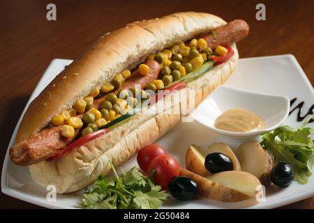Long hot dog avec deux saucisses gros plan, avec des pois verts et du maïs, avec des tranches de tomates et des concombres, sur une assiette blanche avec de la moutarde, des pommes de terre et Banque D'Images