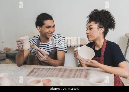 un jeune couple afro-américain vitriant des pots d'argile avec des brosses près de la table avec de l'équipement en poterie Banque D'Images