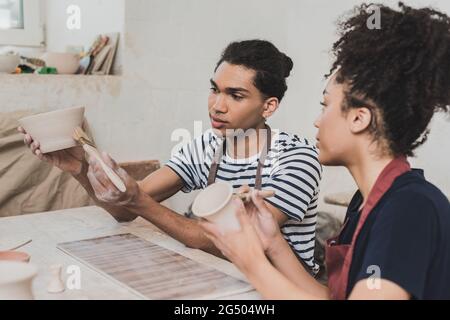 jeune couple afro-américain concentré glaçant des pots d'argile avec des brosses près de la table avec de l'équipement en poterie Banque D'Images