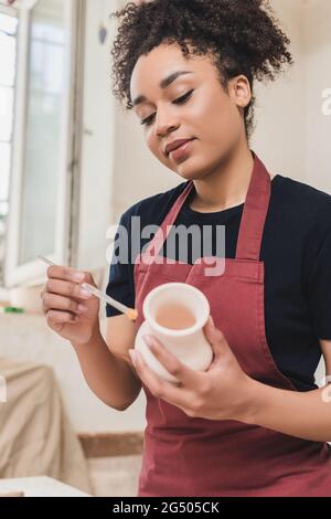 gaie jeune femme afro-américaine vitriant pot d'argile avec brosse en poterie Banque D'Images