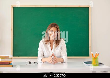 Retour à l'école. Une femme enseignante regarde la caméra tout en étant assise sur un bureau sur le fond d'une commission scolaire dans une classe scolaire. Banque D'Images