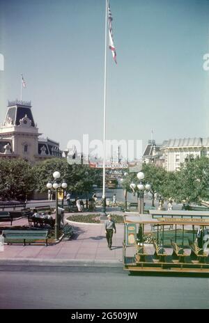 Disneyland, Californie, 1959. Vue sur la place de la ville depuis l'extérieur de la gare de main Street. Un mât de pavillon vole les étoiles et les rayures et le drapeau de la Californie pendant que les visiteurs se détendent sur les bancs et passent. Une voiture de rue tirée par des chevaux Disneyland vous attend en premier plan et main Street et Sleeping Beauty’s Castle sont visibles au loin. Une bannière « Disneyland 59 » est affichée de l'autre côté de main Street, annonçant les nouveaux ajouts du complexe pour cette année. Banque D'Images