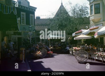 Disneyland, Californie, 1959. Vue sur le marché aux fleurs de main Street sur West Center Street. À droite, les visiteurs sont assis sous des parasols à l'extérieur du salon de la crème glacée Carnation. Sur la gauche se trouve l'auvent de la pharmacie Upjohn. Une bannière « Disneyland 59 » annonçant les nouveaux ajouts de la station pour cette année est affichée sur un lampadaire. Banque D'Images