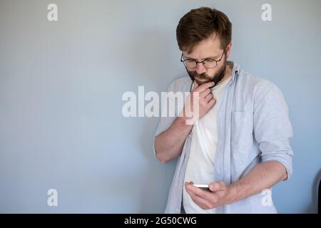 Homme pensif avec des lunettes regarde un smartphone, il est confus et tient le menton avec la main. Copier l'espace. Arrière-plan gris. Banque D'Images