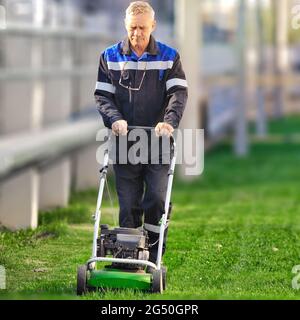 Un travailleur âgé tond la pelouse par temps ensoleillé. Un homme avec des vêtements de travail travaille avec une tondeuse sur un pré vert. Vue avant. Banque D'Images