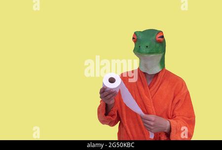 jeune garçon avec un masque d'animal grenouille vert dans un peignoir orange avec un rouleau de papier toilette blanc sur un fond jaune avec espace de copie conceptuel Banque D'Images