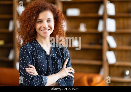 Femme ambitieuse, souriante et murée, debout dans l'espace de bureau avec ses bras croisés et regardant la caméra, charmante étudiante aux cheveux rouges dans la bibliothèque, femme entrepreneure prospère à l'intérieur Banque D'Images