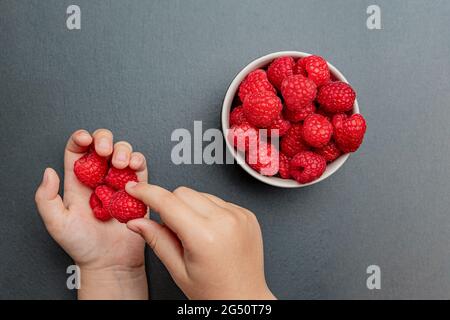 La main de l'enfant prend des framboises juteuses fraîches d'une petite assiette noire. Gros plan rouge vif. Heure d'été de cueillette des baies. Biologique sain Banque D'Images