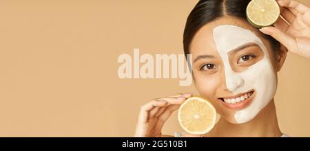 En-tête de site Web du portrait de beauté de la jeune femme avec masque facial appliqué sur la moitié de son visage tenant une tranche de lime et de citron, posant isolé plus Banque D'Images